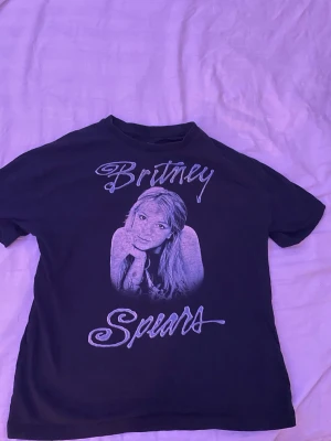 Svart Britney Spears t-shirt - Cool svart t-shirt med stort tryck av Britney Spears på framsidan och hennes namn i vit text. Klassisk passform och rund halsringning. Perfekt för dig som gillar ikoniska musikmotiv och vill sticka ut lite extra.
