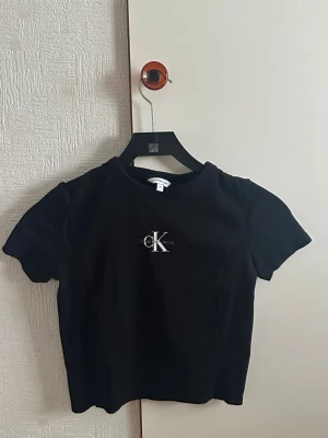 Svart croppad t-shirt Calvin Klein - Snygg svart croppad t-shirt från Calvin Klein med klassisk CK-logga i vitt på bröstet. T-shirten har korta ärmar och rund halsringning. Perfekt till jeans eller kjol för en clean och stilren look. Pris går att diskutera 