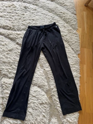 Svarta mjukisbyxor med snörning från Ralph lauren - Svarta mjukisbyxor med resår och snörning i midjan. Byxorna har raka ben och är tillverkade i ett mjukt och bekvämt material, perfekta för att chilla hemma. Fickor på sidorna för extra funktionalitet.