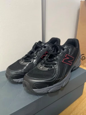 Svarta New Balance sneakers - Snygga svarta sneakers från New Balance med mesh- och syntetmaterial. Skorna har en chunky sula och den klassiska N-loggan i svart med röd kant på sidan. Perfekta för dig som gillar sportig och stilren look. Använd 1 gång 