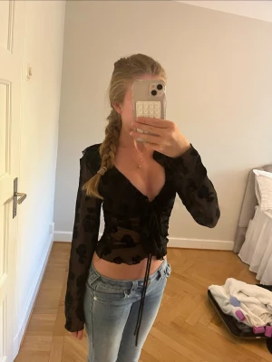 Svart mesh blus med snörning - Trendig svart mesh blus med blommigt mönster och snörning framtill. Blusen är genomskinlig, har v-ringning och långa ärmar. Perfekt att styla med jeans eller kjol för en cool look. Använd en eller två gånger 