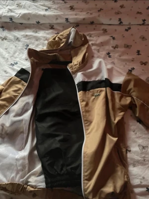 Beige och vit vindjacka  - Snygg vindjacka i beige, vit och svart. Jackan har huva, dragkedja och meshfoder på insidan. Klassisk Lacoste-logga på bröstet och kontrastpaneler över axlarna. Perfekt för en sportig look.