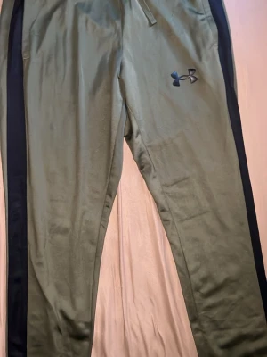 Mörk grön Under Armour - Säljer ett matchande träningsset från Under Armour i olivgrönt med svarta detaljer. Setet består av en zip hoodie och ett par byxor med svarta sidopaneler och logga på både jacka och byxa. Perfekt för gymmet eller löprundan.