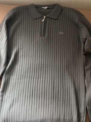 Grå stickad tröja med dragkedja - Snygg grå stickad tröja från Lacoste med diskret logga på bröstet. Tröjan har långa ärmar, ribbade muddar och en liten dragkedja vid kragen. Klassisk pikékrage och vertikalt ribbat mönster ger en stilren look.