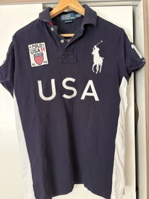Mörkblå vintage piké från Ralph Lauren -  Mörkblå vintage piké/rugbytröja från Polo Ralph Lauren med stora broderade USA-detaljer och Big Pony-logga. Storlek M (Custom Fit). Bra vintage-skick med normalt slitage.  Perfekt för preppy, old money och vintage-stil.
