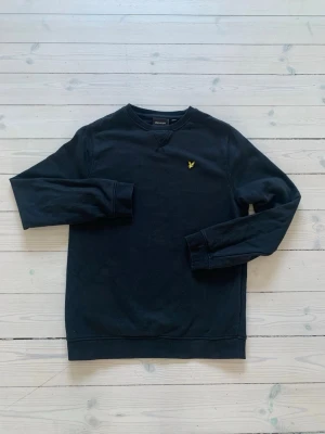 Svart sweatshirt från Lyle & Scott - Klassisk svart sweatshirt från Lyle & Scott med rund halsringning och den ikoniska gula örnloggan broderad på bröstet. Tröjan har långa ärmar, ribbade muddar och är gjord i mjukt bomullsmaterial. Perfekt för en avslappnad och stilren look.