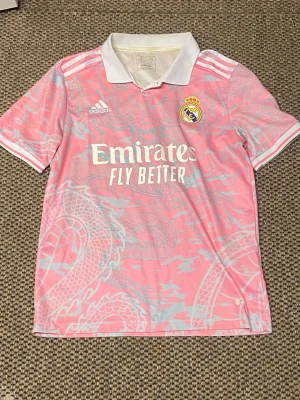 Rosa Real Madrid matchtröja Adidas - Säljer en rosa Real Madrid matchtröja från Adidas med vit krage och detaljer. Tröjan har ett drakmönster i ljusrosa, klubbmärke på bröstet och 'Emirates Fly Better' tryckt framtill. Perfekt för dig som vill sticka ut på planen eller läktaren.