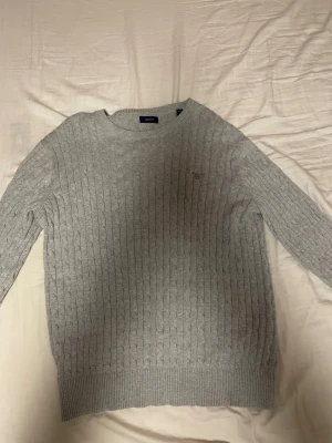 Ljusgrå kabelstickad tröja GANT - Säljer en ljusgrå kabelstickad tröja från GANT med rund halsringning och diskret logga broderad på bröstet. Tröjan har långa ärmar och ribbade muddar vid ärmslut och nederkant. Perfekt för lager på lager och enkel att matcha.
