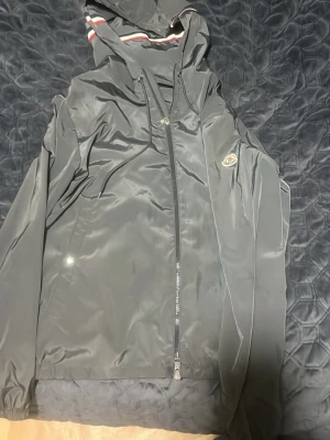 Svart vindjacka från Moncler - Snygg svart vindjacka från Moncler med huva och dragkedja framtill. Jackan har Moncler-logga på ärmen och vita samt röda detaljer på insidan av huvan. Tillverkad i ett glansigt, lätt material som står emot vind. Perfekt för dig som vill ha en stilren och sportig look.