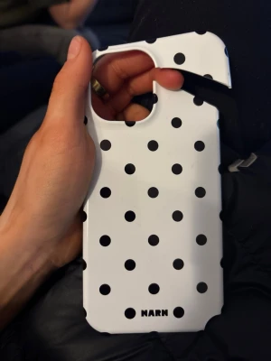 Vitt mobilskal med svarta prickar NARN - TILL IPHONE 14/15 Snyggt mobilskal från NARN i hårdplast med vit bas och svarta prickar. Skalets design är stilren och lekfull, perfekt för dig som vill ha något unikt till mobilen. Passar dig som gillar grafiska mönster och enkel, modern stil.