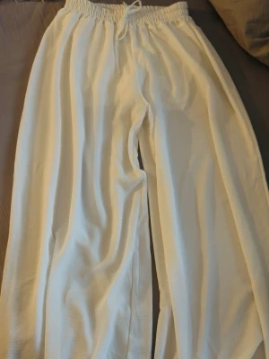 Vita vida mjukisbyxor med dragsko - Säljer ett par vita mjukisbyxor med bred passform och resår i midjan samt dragsko. Byxorna är tillverkade i ett mjukt och bekvämt material som passar perfekt för chill dagar. Inga synliga loggor eller märken.