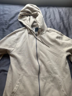 Beige zip hoodie från 157 - Säljer en beige hoodie med dragkedja från 157. Hoodien har huva med dragsko, två stora fickor framtill och är gjord i mjukt sweatshirtmaterial. Perfekt för chill dagar och enkel att matcha med det mesta.