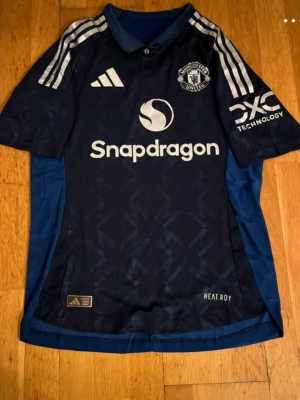 Manchester United bortatröja Adidas - Mörkblå Manchester United bortatröja från Adidas med vita detaljer och loggor. Tröjan har Snapdragons sponsortryck på bröstet, klassiska Adidas-ränder på axlarna och klubbmärke på vänster sida. Tillverkad i lätt och ventilerande material.