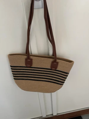 Beige stråväska med bruna handtag - Snygg beige handväska i stråmaterial med svarta dekorativa ränder och långa bruna handtag i konstläder. Väskan har en rymlig form och passar perfekt för dig som gillar en avslappnad men trendig stil. Perfekt accessoar till sommaren!