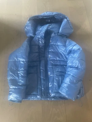 Blå glansig pufferjacka med huva - Snygg blå pufferjacka med glansigt material och stor huva. Jackan har två stora fickor framtill och dragkedja med tryckknappar. Perfekt för kalla vinterdagar och ger en cool streetwear-look.