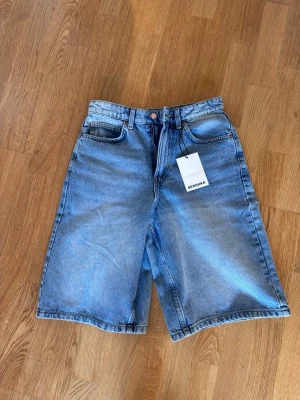 Blå denimshorts från Bershka - Säljer ett par blå denimshorts från Bershka i klassisk femficksmodell. Shortsen har hög midja, raka ben och en avslappnad passform. Tillverkade i bomull med en ljus tvättad look och knappgylf. Perfekta för sommaren!