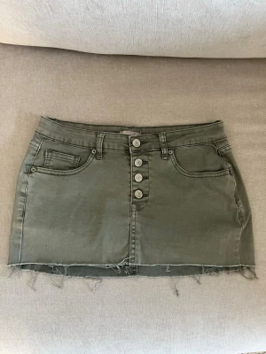 Olivgrön kort jeanskjol - Snygg olivgrön kort jeanskjol med rå kant och fyra silverfärgade knappar framtill. Kjolen har klassiska fickor både fram och bak samt hällor för bälte. Skriv för mått