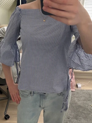 Blårandig offshoulder blus - Snygg blårandig offshoulder-blus med trekvartsärmar och volangdetaljer samt knytband vid ärmarna. Tunn och luftig modell i bomullsmix, perfekt för en trendig och avslappnad look. Klassiska smala ränder i vitt och blått.