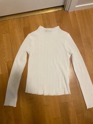 Vit ribbad långärmad tröja - En stilren vit långärmad tröja med ribbad struktur och hög krage. Perfekt att matcha med jeans eller kjol för en clean look. Materialet är mjukt och stretchigt, vilket gör den bekväm att bära hela dagen.