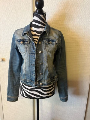 s. Oliver Blå klassisk jeansjacka - Snygg blå jeansjacka med knappar framtill och två bröstfickor med lock. Jackan har en klassisk krage och långa ärmar med knapp vid ärmslut. Perfekt för att ge din outfit en cool touch. Materialet är slitstarkt denim och jackan har en figurnära passform.