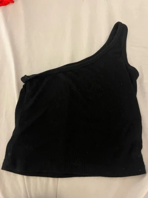 Svart oneshoulder croppad topp - Säljer en svart oneshoulder topp som är croppad och har ribbad struktur. Perfekt för dig som gillar en enkel men edgy stil. Materialet är mjukt och stretchigt, vilket gör den bekväm att bära. Passar till många olika outfits.