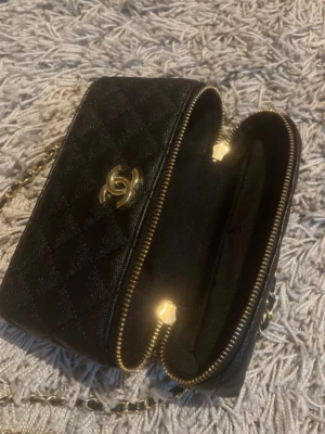 Svart väska - Säljer en svart quiltad handväska med klassiskt guld CC-logo framtill. Väskan har kedjerem med läderdetaljer och guldfärgade dragkedjor. Materialet är läder med struktur och diamantmönster. Perfekt för dig som vill ha en lyxig och tidlös accessoar.