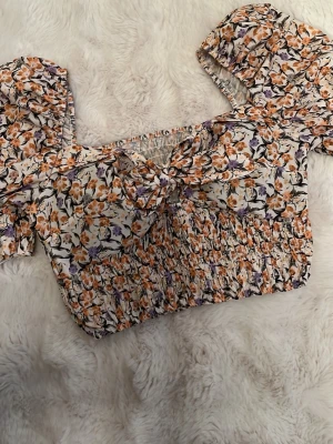 Blommig croppad blus med puffärm - Supersöt magtröja med blommigt mönster i orange, lila och svart på vit botten. Går att använda som offshoulder och vanligt. Toppen har puffiga korta ärmar, smockad midja och en söt knytning framtill. Perfekt för sommarens alla tillfällen!