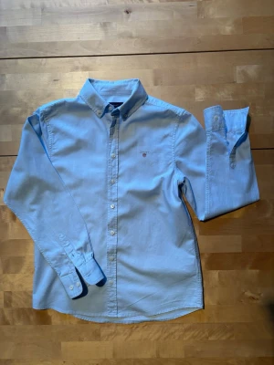 Ljusblå skjorta från Gant - Klassisk ljusblå skjorta från Gant med button-down krage och broderad logga på bröstet. Skjortan har långa ärmar, knappar framtill och är tillverkad i mjuk bomull. Perfekt för en clean och stilren look.