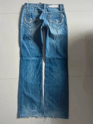 Blå bootcut jeans  - Säljer ett par blå bootcut jeans från True Religion med klassiska kontrastsömmar och broderade fickor bak. Jeansen har låg midja, slitningar och rå kant nertill för en avslappnad look. Perfekta för dig som gillar en vintage vibe och vill sticka ut.