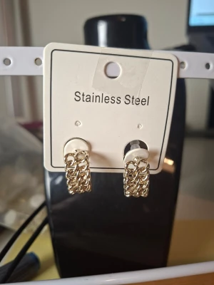 Guldiga kedjeörhängen i rostfritt stål - Säljer ett par snygga örhängen i rostfritt stål med guldfinish. Örhängena har en trendig kedjedesign och är halvrunda hoops som ger en cool och modern look. Perfekta för dig som gillar statement-accessoarer och vill sticka ut lite extra.