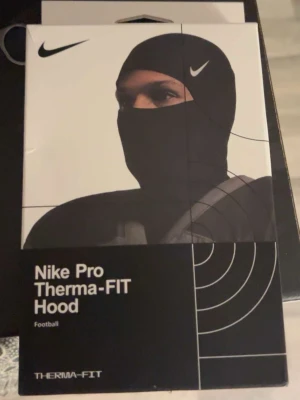 Nike Pro Therma-FIT Hood - Nike Pro Therma-FIT Hood är en svart huva designad för att hålla dig varm under träning eller fotboll. Den är tillverkad i ett tekniskt syntetmaterial som hjälper till att reglera värmen och har en tight passform som täcker huvud och hals.