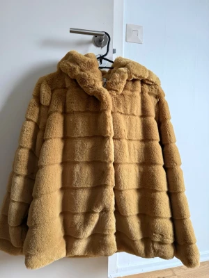 Camelbrun fuskpälsjacka Gold & Silver - Säljer en fluffig camelbrun fuskpälsjacka från Gold & Silver. Jackan har huva, breda horisontella paneler och stängs framtill. Perfekt för dig som vill ha en varm och trendig look under kalla dagar.