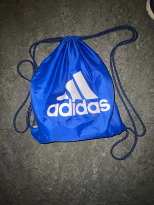 Blå gympåse från Adidas - Säljer en blå gympåse från Adidas med stort vitt logotryck på framsidan. Väskan har dragskostängning och tunna snören som axelband. Perfekt för träning eller att bära med sig det viktigaste. Tillverkad i lätt och slitstarkt material.