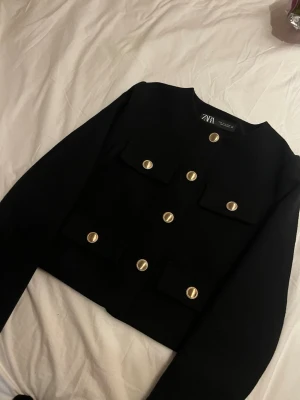 Svart kavajkofta från ZARA - Stilren svart kavajkofta från ZARA med rund halsringning och dekorativa guldknappar framtill. Fyra fickor med lock ger en trendig look. Långärmad och i ett tjockare tyg som känns lyxigt. Perfekt för dig som gillar en chic och modern stil.