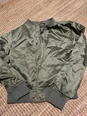 Olivgrön bomberjacka XS Black Label - Snygg olivgrön bomberjacka från The Black Label Collection i storlek XS. Jackan har klassisk ribbad mudd vid ärmslut och midja, dragkedja framtill och en stilren look. Tillverkad i glansigt material som ger en modern vibe.