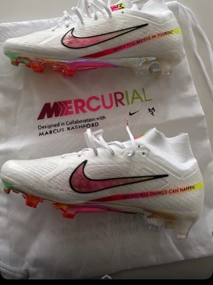 Nike Mercurial fotbollsskor Marcus Rashford - Säljer ett par vita Nike Mercurial fotbollsskor i samarbete med Marcus Rashford. Skorna har regnbågsfärgad sula, svart och röd swoosh samt texten 'When you believe in yourself' och 'Incredible things can happen' på sidan. Lätta och ventilerande material för bästa prestanda.