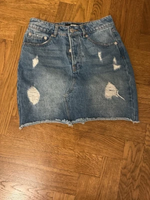 Blå jeanskjol från BikBok - Säljer en blå jeanskjol från BikBok i storlek XS. Kjolen har slitna detaljer, rå kant nertill och klassisk femficksmodell. Perfekt för dig som gillar en avslappnad och trendig stil.