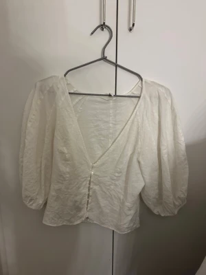 Vit puffärmad blus med knappar - Trendig vit blus med puffiga trekvartsärmar och djup v-ringning framtill. Blusen har knäppning med små pärlemorknappar och är tillverkad i ett lätt, krispigt tyg som ger en luftig känsla. Perfekt för en stilren och modern look. Från hm i storlek S