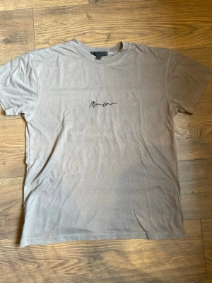 Beige t-shirt från Mennace - Säljer en beige t-shirt från Mennace med svart broderad logga på bröstet. Klassisk rund och lite tjockare halsringning med korta ärmar. Tillverkad i mjuk bomull för skön känsla och enkel stil.