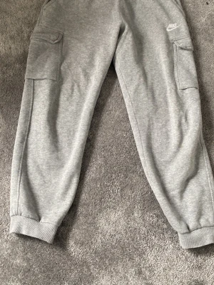 Nike byxor - Säljer ett par gråa mjukisbyxor från Nike med cargo-fickor på sidorna och resår i midjan samt vid bensluten. Byxorna har dragsko i midjan och är tillverkade i mjukt sweatshirtmaterial. Perfekta för en avslappnad och sportig stil.