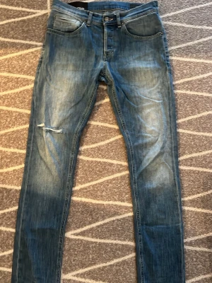 Blå slitna jeans från Dondup - Säljer ett par blå jeans från Dondup med skinny fit och slitningar på vänster knä. Jeansen har klassiska fem fickor, orangea sömmar och en cool tvättad look. Perfekta för dig som gillar en avslappnad och trendig stil.
