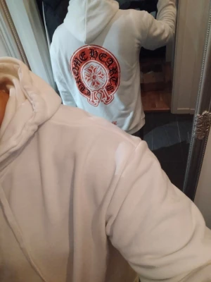 Vit Chrome Hearts hoodie med tryck - Säljer en vit hoodie från Chrome Hearts med stort rött och svart tryck på ryggen och mindre tryck på bröstet. Hoodien har huva med dragsko och en klassisk känguruficka framtill. Perfekt för dig som gillar streetwear och snygga detaljer.