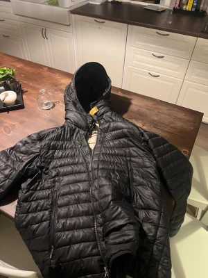 Parajumper jacka - Snygg svart dunjacka från Parajumpers, modell Day Tripper. Jackan har huva, dragkedja framtill och två stora fickor med dragkedja. Lättviktigt material med quiltad design och cool detalj med gul stropp vid halsen. Perfekt för våren och sena sommarkvällar