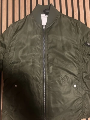 Olivgrön bomberjacka från Kingfield - Snygg olivgrön bomberjacka från hope med klassisk ribbad krage och silverfärgad dragkedja. Jackan har två fickor med tryckknappar och en ärmficka med dragkedja. Materialet är glansigt och lätt, perfekt för att ge en cool look. Passar M till S. Den är puffig och passar en avslappnad stil.