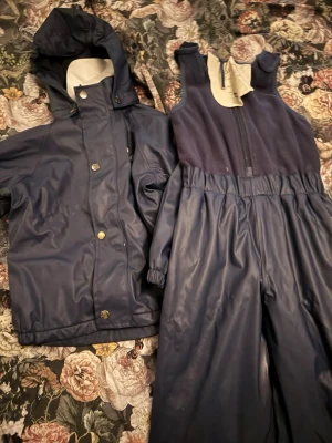 Mörkblå regnbyxor o regnjacka (fodrad) - Säljer ett sett mörkblå regnkläder (kuling) gjorda i ett vattenavvisande material, perfekta för kalla och blöta dagar. Matchande jacka med huva ingår. Storlek 110/116
