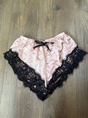 Rosa satinshorts med spetsdetaljer - Supersöta pyjamasshorts i ljusrosa satin med breda svarta spetskanter och en liten svart rosett framtill. Shortsen har elastisk midja och mjukt, glansigt material som känns lyxigt mot huden. Perfekta för mysiga hemmakvällar.