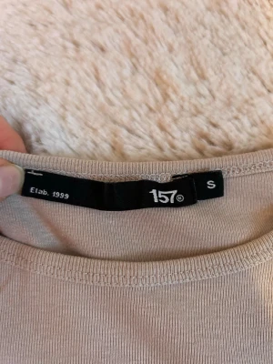Beige långärmad topp från 157 - En basic beige långärmad topp från 157 med rund halsringning. Tillverkad i mjukt material som känns skönt mot huden. Perfekt att styla med jeans eller kjol för en clean och enkel look.