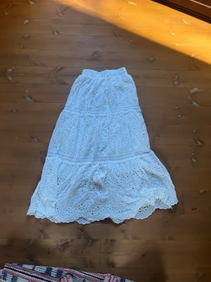 Vit långkjol med broderade detaljer - Säljer en romantisk vit långkjol med broderade hålmönster och volangkant. Kjolen har snörning i midjan med tofsar och är tillverkad i ett lätt bomullstyg, perfekt för varma dagar. Passar dig som gillar boho-stil och vill ha en luftig kjol till sommaren. Köpt i Spanien och aldrig använt 😊 skriv om ni har frågor! 