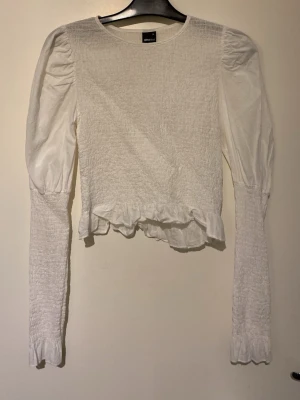 Vit långärmad topp från Gina Tricot - Säljer en vit långärmad topp från Gina Tricot med puffärmar och smockad struktur. Toppen har volangkant nedtill och är croppad i modellen. Perfekt att styla med höga jeans eller kjol för en trendig look.