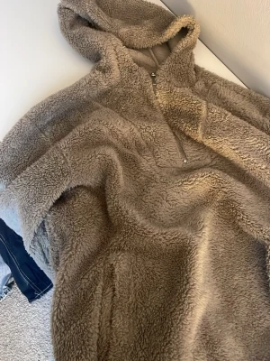 Beige teddyjacka med huva - Supermjuk teddyjacka i beige med dragkedja framtill och stor huva. Jackan har lång ärm och är gjord i fluffigt, värmande material som känns riktigt cozy. Perfekt för kyliga dagar när du vill vara både varm och snygg.
